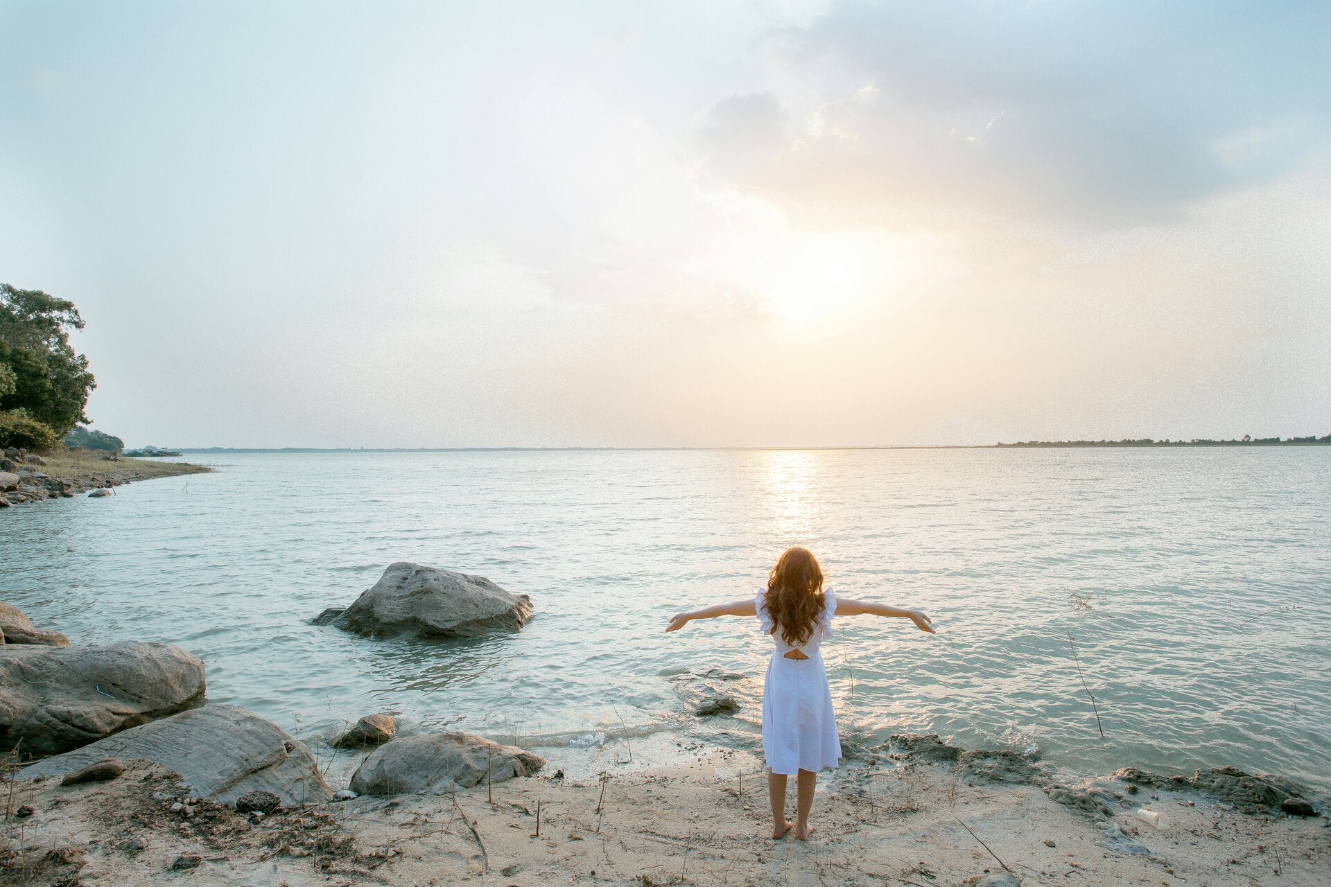 Vrouw in witte jurk staat met gespreide armen aan het water bij zonsondergang